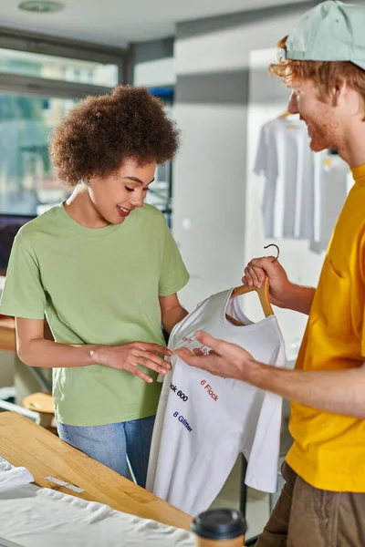 Smiling young african american craftswoman looking at t-shirt with lettering on hanger while colleague talking and working on project in blurred print studio, start-up innovation concept — Stock Photo