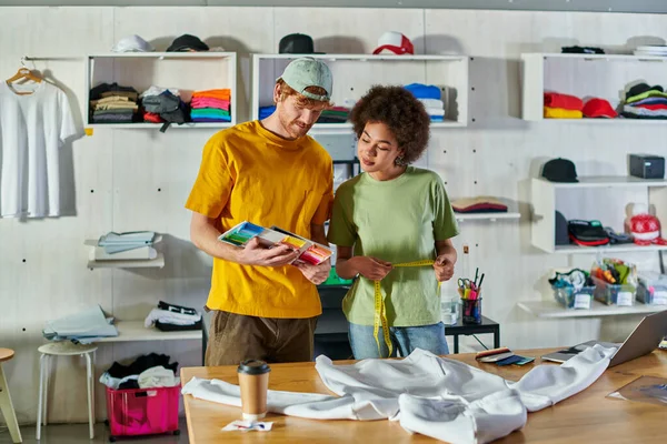 Young craftsman holding cloth samples near african american colleague with tape measure and working with clothes near laptop in print studio, self-employment opportunity concept — Stock Photo