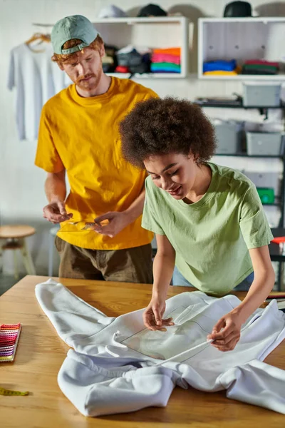 Молодой африканский американский дизайнер держит печатный слой рядом с одеждой и коллегой, работая вместе в размытой типографии на заднем плане, концепция возможности самозанятости — стоковое фото