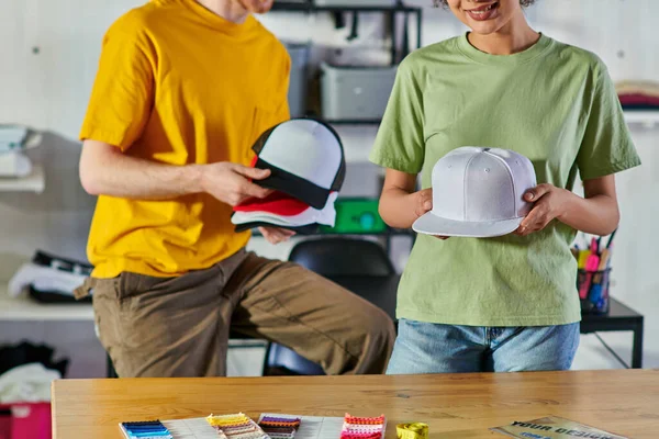 Cropped view of smiling young african american craftswoman holding snapback and working with colleague near cloth swatches in print studio, sustainable business model concept — Stock Photo