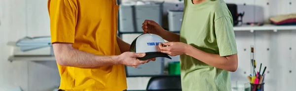 Cropped view of young multiethnic craftspeople holding printing layer near snapback while working in blurred print studio at background, sustainable business model concept, banner — Stock Photo