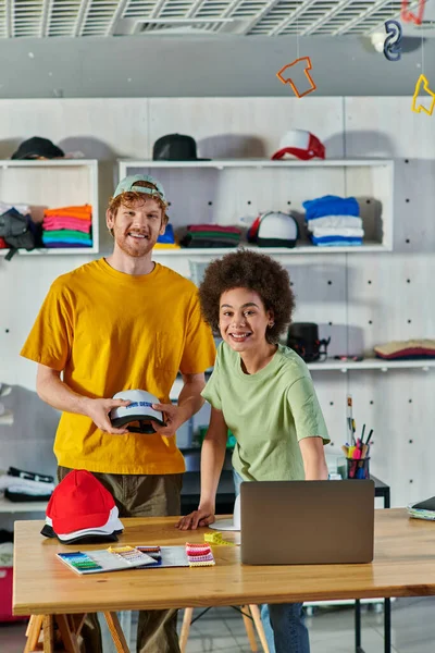 Cheerful young multiethnic designers looking at camera while working with snapbacks, laptop and cloth samples on table in print studio at background, sustainable business model concept — Stock Photo