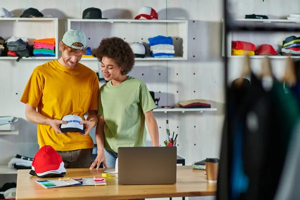 Smiling interracial craftspeople holding snapback and printing layer while working on project near laptop and cloth samples on table in print studio, sustainable business model concept — Stock Photo