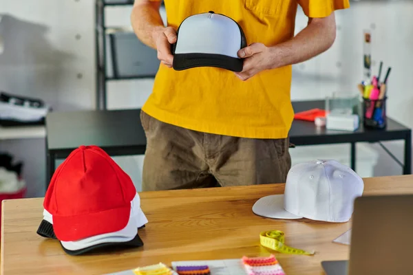 Cropped view of young craftsman holding snapback while working near tape measure and blurred laptop on table in print studio at background, small business resilience concept — Stock Photo