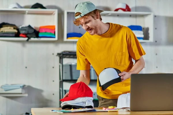 Jovem artesão ruiva alegre segurando snapbacks enquanto trabalhava perto de amostras de pano e laptop borrado na mesa de madeira no estúdio de impressão, conceito de resiliência de pequenas empresas — Fotografia de Stock