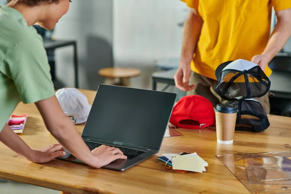 Cropped view of young african american designer using laptop while working near color swatches, coffee to go and colleague with snapback in print studio, small business resilience concept — Stock Photo