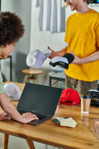 Young african american craftswoman using laptop while working on project near color samples and blurred colleague with snapbacks in print studio, small business resilience concept — Stock Photo