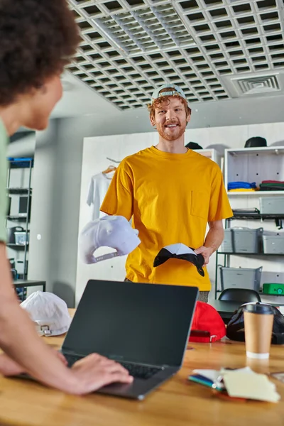 Cheerful young craftsman holding snapbacks and talking to blurred african american colleague using laptop and working on project in print studio, small business resilience concept — Stock Photo