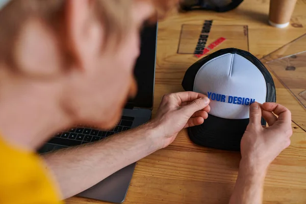 Blurred young designer holding printing layer near snapback while working on project near laptop on table in print studio, hands-on entrepreneurship concept, your design letters — Stock Photo