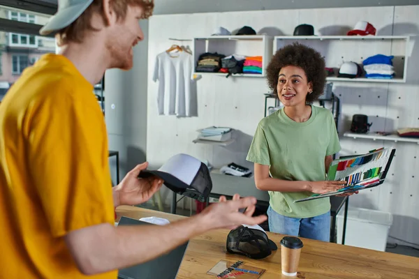 Smiling young african american designer holding cloth samples near blurred colleague with snapback and laptop with blank screen in print studio, hands-on entrepreneurship concept — Stock Photo