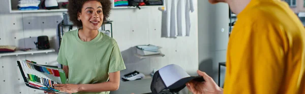 Joven diseñador afroamericano alegre sosteniendo muestras de color y mirando al colega con snapback mientras trabaja en un estudio de impresión borrosa, concepto de emprendimiento práctico, banner - foto de stock