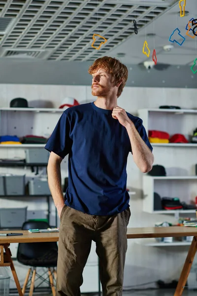 Junger rothaariger Handwerker in lässiger Kleidung, der die Hand in der Tasche hält und wegschaut, während er im verschwommenen Druckstudio steht, praktisches Unternehmertum-Konzept — Stockfoto