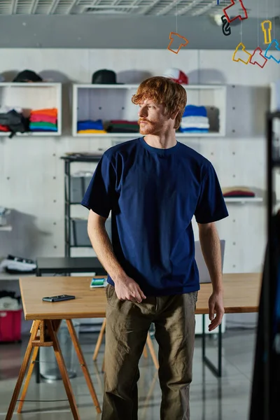 Junger rothaariger Handwerker in lässiger Kleidung, der wegschaut, während er am Arbeitstisch in einem verschwommenen Druckeratelier steht, praktisches Unternehmertum-Konzept — Stockfoto