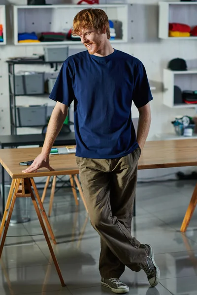 Sonriente joven pelirrojo artesano en ropa casual cogido de la mano en el bolsillo mientras está de pie cerca de la mesa de trabajo en el estudio de impresión, concepto de emprendimiento práctico - foto de stock