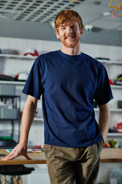 Porträt eines fröhlichen rothaarigen Handwerkers in lässiger Kleidung, der die Hand in der Tasche hält und in die Kamera lächelt, während er im Druckatelier steht, unternehmerisches Konzept zum Anfassen — Stockfoto
