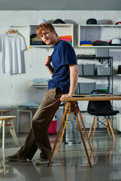 Voller Länge junger rothaariger Handwerker in lässiger Kleidung, der in die Kamera blickt, während er am Smartphone-Arbeitstisch im Druckstudio steht, selbstgemachtes Erfolgskonzept — Stockfoto