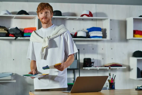 Young redhead craftsman in casual clothes holding cloth samples and looking at camera while standing near laptop on table in blurred print studio, self-made success concept — Stock Photo