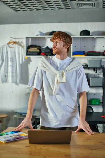 Young redhead craftsman in casual clothes looking away while standing near laptop and cloth samples on table in blurred print studio, self-made success concept — Stock Photo