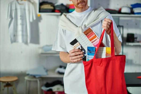 Cropped view of young craftsman in casual clothes putting cloth samples in shoulder bag while working in blurred print studio at background, self-made success concept — Stock Photo
