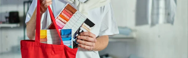Cropped view of young craftsman in casual clothes putting cloth samples in shoulder bag in blurred print studio at background, self-made success concept, banner — Stock Photo