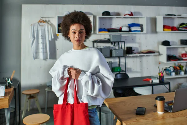 Young african american craftswoman holding shoulder bag and looking at camera while standing near devices and coffee to go on table in print studio, enthusiastic business owner working in workshop — Stock Photo