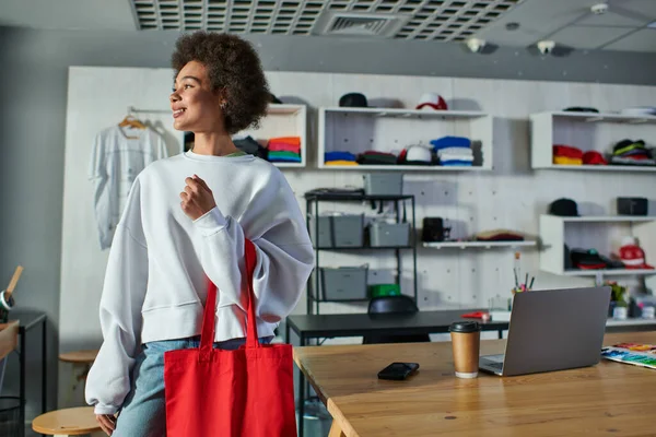 Улыбающаяся молодая африканская американская ремесленница, держащая плечевую сумку и глядя в сторону, стоящая рядом с устройствами и кофе, чтобы пойти в типографию, восторженный владелец бизнеса, работающий в мастерской — стоковое фото