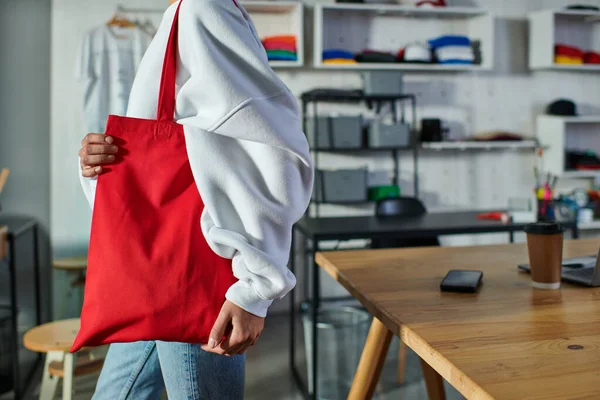 Cropped view of young african american craftswoman holding shoulder bag and standing near blurred devices and coffee to go in print studio, enthusiastic business owner working in workshop — Stock Photo
