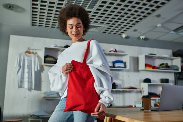 Fröhliche junge afrikanisch-amerikanische Handwerkerin blickt auf Umhängetasche, während sie neben Coffee to go und Laptop auf dem Tisch im Druckstudio steht, begeisterte Geschäftsinhaberin arbeitet in Werkstatt — Stockfoto
