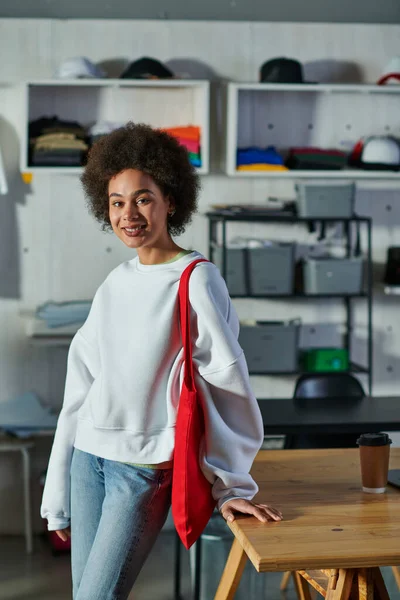 Joven y positiva artesana afroamericana con bolso de hombro mirando a la cámara y de pie cerca del café para ir a la mesa en el estudio de impresión, entusiasta propietario de negocios que trabaja en el taller — Stock Photo