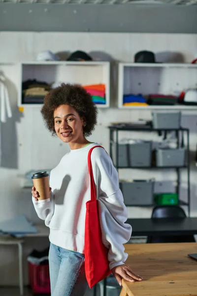 Sorrindo jovem afro-americana artesão com bolsa de ombro segurando café para ir e olhando para a câmera perto da mesa em estúdio de impressão borrada, proprietário de negócios entusiasmado trabalhando em oficina — Fotografia de Stock