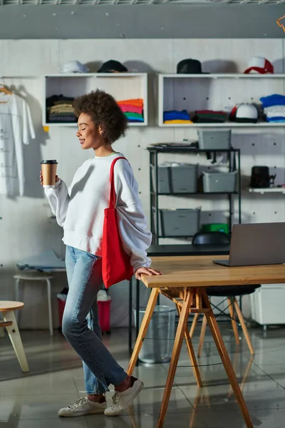 Вид сбоку на молодую и позитивную африканскую сумасшедшую женщину с сумкой на плече, держащую в руках кофе и стоящую рядом с ноутбуком на столе в типографии, увлеченного владельца бизнеса, работающего в мастерской — стоковое фото