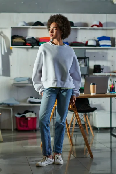 Повна довжина молодої афроамериканської майстрині в сорочці і джинсах, дивлячись далеко, стоячи біля робочого столу в друкованій студії, захоплений власник бізнесу працює в майстерні — стокове фото