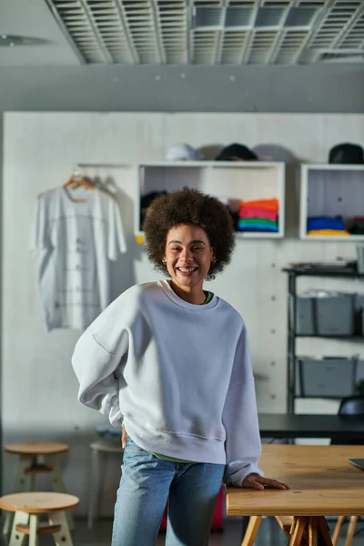 Веселая и уверенная в себе африканская ремесленница в толстовке и джинсах, смотрящая в камеру, стоя возле рабочего стола в типографии, восторженный владелец бизнеса, работающий в мастерской — стоковое фото