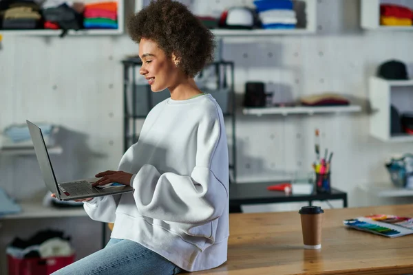 Jovem artesão afro-americana positiva em jeans e camisola usando laptop enquanto se senta perto de café para ir à mesa no estúdio de impressão, proprietário de negócios entusiasmado trabalhando em oficina — Fotografia de Stock