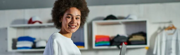 Jovem trabalhadora afro-americana alegre em camisola olhando para a câmera perto do laptop em estúdio de impressão borrada em segundo plano, focada em oficina de gerenciamento de proprietário de negócios, banner — Fotografia de Stock
