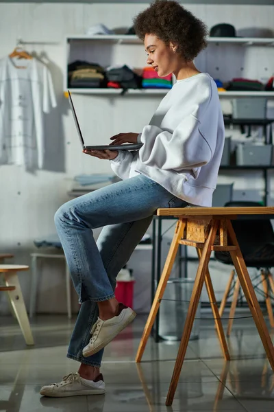 Junge afrikanisch-amerikanische Handwerkerin in Jeans und Sweatshirt mit Laptop und auf dem Tisch sitzend, während sie im verschwommenen Druckereistudio arbeitet, fokussierte Geschäftsinhaberin, die die Werkstatt leitet — Stockfoto