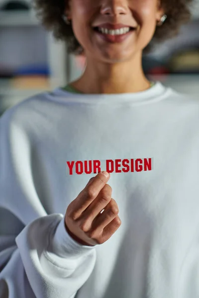 Ausgeschnittene Ansicht eines lächelnden verschwommenen afrikanisch-amerikanischen Designers in Sweatshirt, der Druckschicht mit Ihrem Design-Schriftzug im Druckstudio hält, fokussierter Geschäftsinhaber, der die Werkstatt leitet — Stockfoto