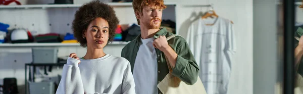Confident african american artisan looking at camera while standing near redhead colleague with bag in blurred print studio, collaborative business owners working together, banner — Stock Photo