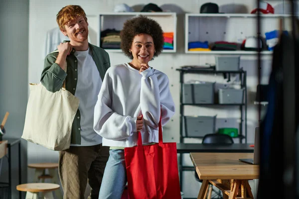 Smiling young african american artisan holding shoulder bag and looking at camera near redhead colleague in blurred print studio, collaborative business owners working together — Stock Photo
