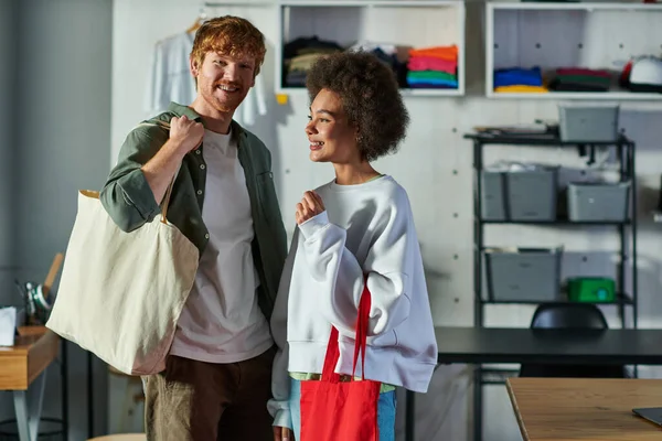 Smiling redhead craftsman holding shoulder bag and looking at camera near african american colleague while standing in print studio, collaborative business owners working together — Stock Photo