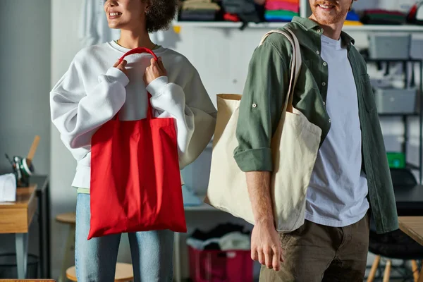 Cropped view of positive young african american craftswoman holding bag and standing near colleague in blurred print studio, collaborative business owners working together — Stock Photo