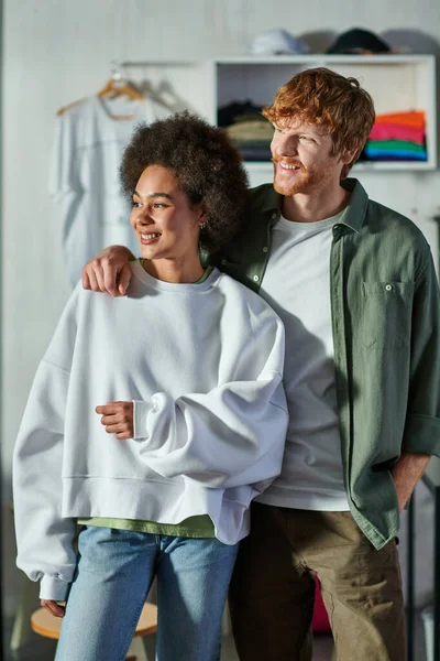 Lächelnder rothaariger Handwerker umarmt afrikanisch-amerikanischen Kollegen und schaut weg, während er im verschwommenen Druckstudio im Hintergrund steht, kooperative Geschäftsinhaber arbeiten zusammen — Stockfoto