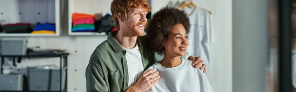 Cheerful young redhead craftsman hugging african american colleague and looking away while standing in print studio, collaborative business owners working together, banner — Stock Photo