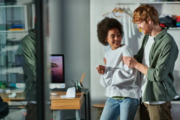 Alegre pelirroja artesano abrazando africano americano colega y hablando mientras de pie en borrosa impresión studio, colaborativo negocios propietarios trabajando juntos, divertirse - foto de stock