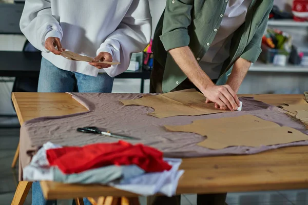 Vista cortada de artesã afro-americana segurando padrão de costura enquanto colega marcando tecido na mesa em estúdio de impressão, empresários colaborativos trabalhando juntos — Fotografia de Stock