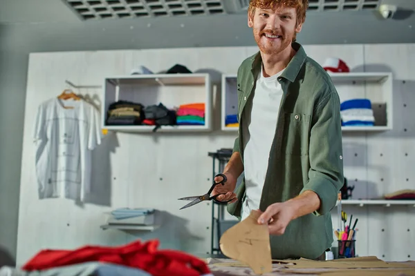 Lächelnder rothaariger Handwerker, der in die Kamera schaut, während er Schere und Schnittmuster in Stoffnähe auf dem Tisch im Druckstudio hält, Multitasking-Unternehmer, der mehrere Projekte verwaltet — Stockfoto