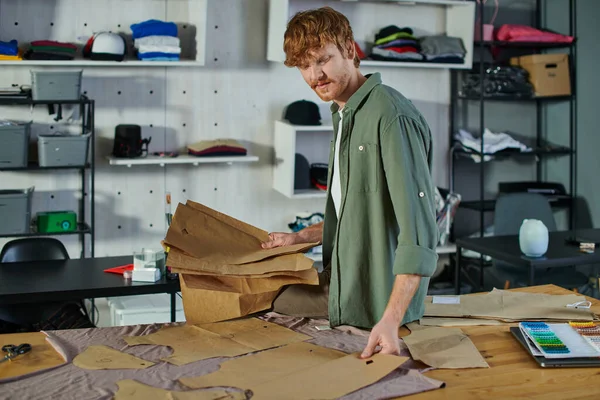 Junger rothaariger Handwerker, der Schnittmuster auf Stoff legt, während er in der Nähe von Scheren und Stoffmustern auf dem Tisch im Druckstudio arbeitet, Multitasking-Unternehmer, der mehrere Projekte verwaltet — Stockfoto