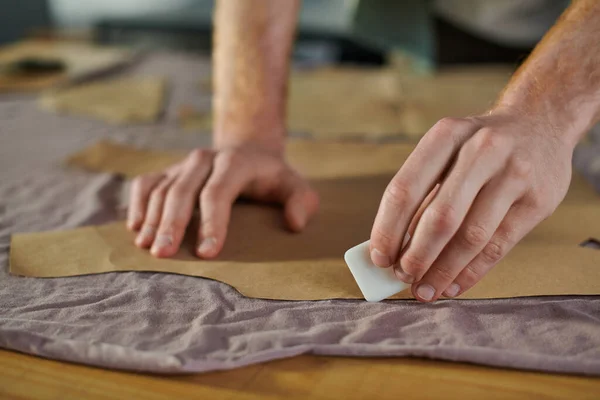 Ausgeschnittene Ansicht eines jungen Handwerkers, der Stoff mit Kreide markiert, während er mit Schnittmustern auf dem Tisch im Druckstudio arbeitet, Multitasking-Unternehmer, der mehrere Projekte verwaltet — Stockfoto