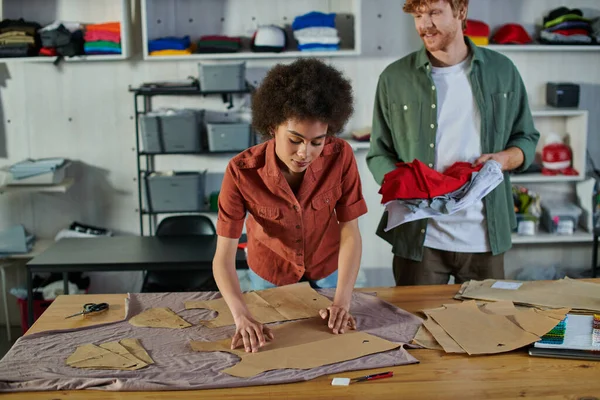 Jovem artesã afro-americana colocando padrões de costura em tecido e trabalhando perto de sorrir colega ruiva no estúdio de impressão, ambicioso conceito de jovens empreendedores — Fotografia de Stock