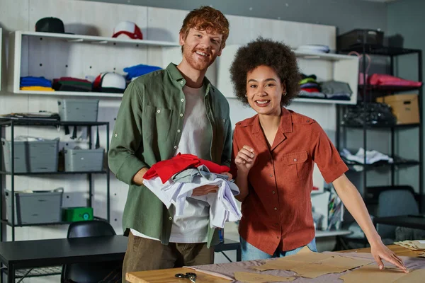 Positive young multiethnic craftspeople looking at camera while working with clothes, fabric and sewing patterns in print studio at background, ambitious young entrepreneurs concept — Stock Photo